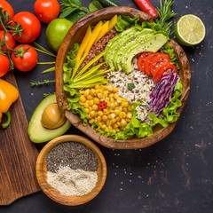 Buddha bowl with chickpea, avocado, wild rice, quinoa seeds, bell pepper, tomatoes, greens, cabbage, lettuce on shabby dark stone table and wooden bowl with chia and sesame seeds. Top view.