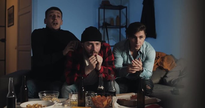 Three friends praying while watching football match on television at home, cheering best soccer team. Emotion. Men's fans celebrate winning.