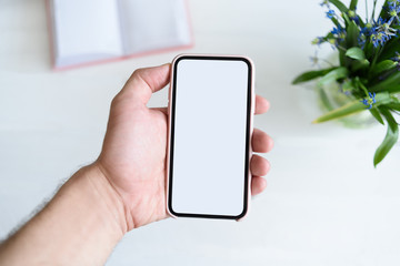 Male hand with a smartphone. White blank screen. Table with notebook and flowers on background