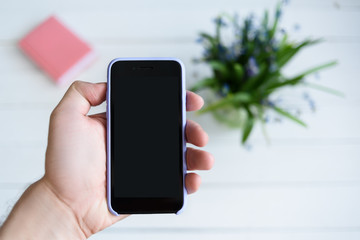 Male hand with a smartphone. Black blank screen. Table with notebook and flowers on background