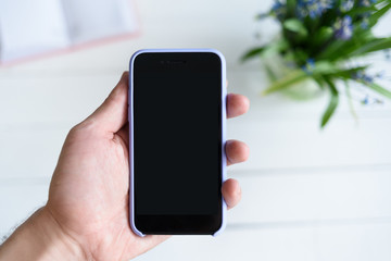 Male hand with a smartphone. Black blank screen. Table with notebook and flowers on background