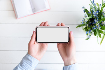 Female hands with a smartphone. White blank screen. Table with notebook and flowers on background