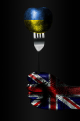 A hand with a drawn Great Britain flag holds a fork, on which is a ball with a drawn Ukraine flag,...
