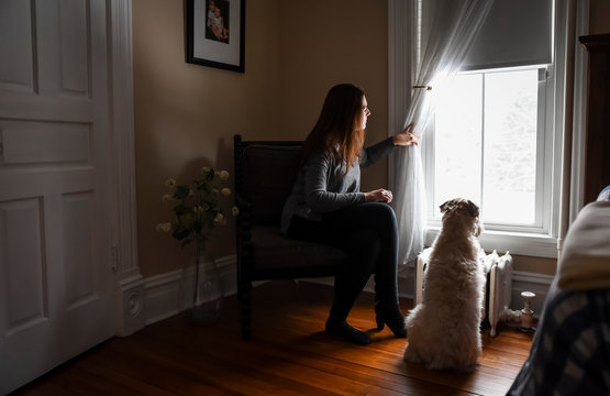 Woman Pulling Back Curtain To Look Out Window With Dog.