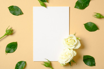 Blank white paper card and frame made of roses flowers and green leaves on pastel yellow background. Top view, tender minimal flat lay style composition. Creative layout, flower arrangement concept.