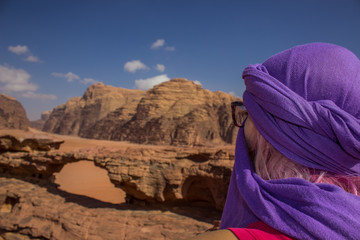 traveler woman in hijab Arabian traditional cloth on head back to camera looking side ways on bridge shape heritage natural rocky object in Jordanian Wadi Rum desert scenic environment