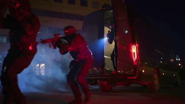 Masked Team of Armed SWAT Police Officers Exit a Black Van Parked Outside of an Office Building. Soldiers with Rifles and Flashlights Run on a Street Filled with Smoke.