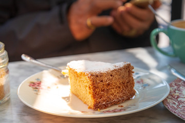 Close up view on a piece of cake with icing sugar, on the white plate beside sugar bottle and cup of coffee, and outdoor vintage sunshine atmosphere with blur background people use mobile phone.