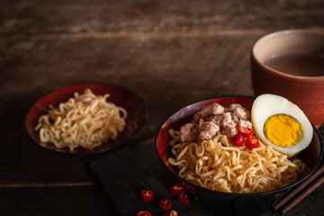 Instant noodle with pork, egg and vegetables on black bowl on the wood table there are chilli, chopstick, instant noodle in black plate and drinking glass placed around.