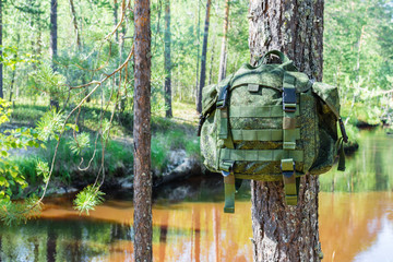 Small green travel backpack hangs on tree branch against river in forest.