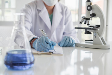 A scientist hands writing on a clipboard in laboratory with test tube microscope and solutions.