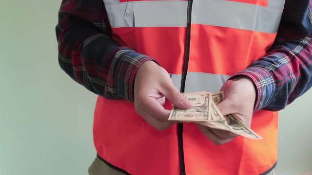 A working man wearing a reflective vest counts ten-dollar bills and give it to the camera. Hands closeup isolated on light green background.
