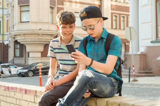 Outdoor Portrait Of Two Boys 13, 14 Years Old, Talking On City Street, Friends Laughing, Looking At Mobile Phone