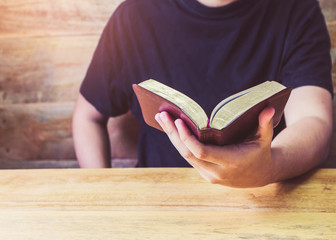 Close up of young man hands holding holy bible on wooden table over wood wall background, Christian...
