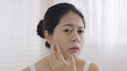 Photo upset asian woman squeezing pimple on face while standing on white background copy space. young elegant wife resting at home looking camera as mirror checking cheek frowning unhappy dry skin