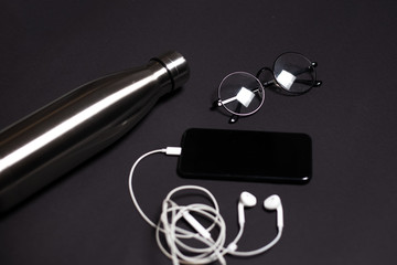 Top view of black desk with steel thermo bottle, glasses, smartphone and earphones.