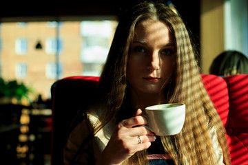 Young pretty white girl in casual clothing sits on a red couch and drinks coffee near a window in a cafe. Shade from the blinds. Thin lines of light on the hair.