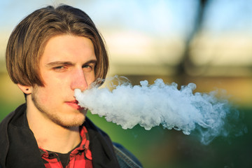 Vape teenager. Portrait of young handsome guy smoking an electronic cigarette  outdoors in a park in spring. Bad habit that is harmful to health. Vaping activity.