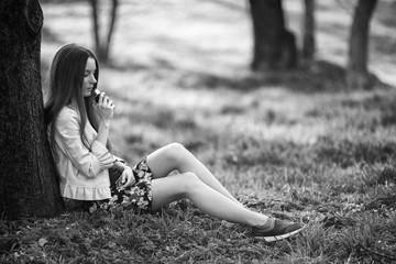 Vape teenager. Young cute girl in a dress sits near a tree and smokes an electronic cigarette outdoors in a park in spring. Bad habit that is harmful to health. Vaping activity. Black and white.