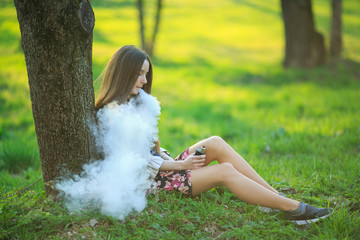 Vape teenager. Young cute girl in a dress sits near a tree and smokes an electronic cigarette...