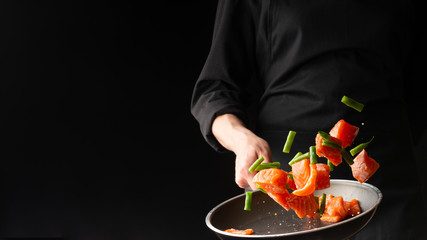 Chef prepares pieces of salmon or trout fillet with green beans in a pan, on a black background for design, recipe book, menu, restaurant or hotel sign, cooking, gastronomy