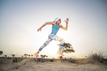 Side view of a strong African Black woman with long braids in hair in the desert in sportswear does a dramatic running leap against a golden sunset or sunrise  holding a bamboo stick   