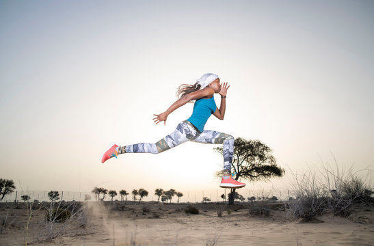 Side view of a strong African Black woman with long braids in hair in the desert in sportswear does a dramatic running leap against a golden sunset or sunrise  holding a bamboo stick   