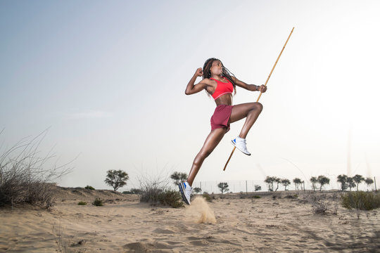 Side view of a strong African Black woman with long braids in hair in the middle eastern desert in sportswear does a dramatic running leap against a golden sunset or sunrise holding a bamboo stick - Powered by Adobe