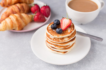 A stack of pancakes with berries, croissant and cup of coffee on gray background