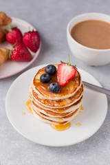 A stack of pancakes with berries and honey, croissant and cup of coffee on gray background