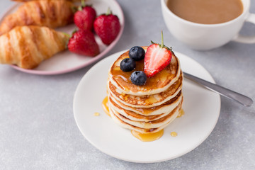 A stack of pancakes with berries honey, croissant and cup of coffee on gray background