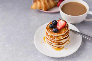 A stack of pancakes with berries, honey, croissant and cup of coffee on gray background