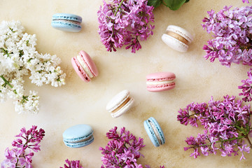 Fototapeta na wymiar Minimalistic composition with bunch of french macaron sweets of different color and taste, lilac flowerings scattered over yellow concrete texture background. Top view, close up, flat lay, copy space.