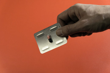 Male hand is holding a medicinal pill. The capsule in a blister. Red background. Concept disease and treatment, medicine and health.