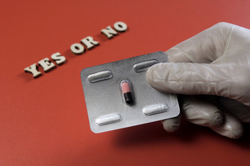 Male hand in white glove holds a medicinal pill. The inscription YES OR NO from wooden letters. The capsule in a blister. Red background. Concept of choice, illness and treatment, medicine and health.