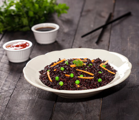 Cooked black rice with vegetables in a plate with sauces and chopsticks on the wooden background.