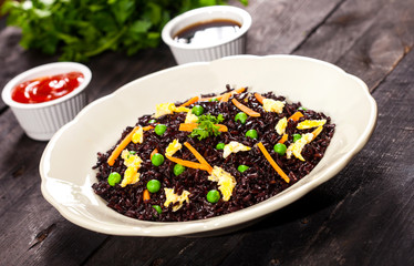 Cooked black rice with vegetables in a plate with sauces and chopsticks on the wooden background.
