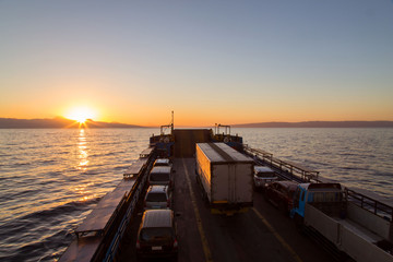 Fototapeta na wymiar Ferry car and truck transportation in an idyllic sunset back lighted tranquil scene with orange reflection in water and sunbeams 