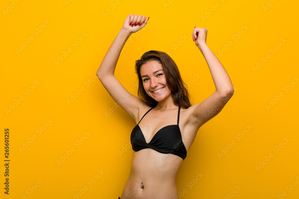 Wall mural Young european woman wearing bikini celebrating a special day, jumps and raise her arms with energy.