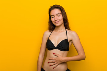 Young european woman wearing bikini touches tummy, smiles gently, eating and satisfaction concept.
