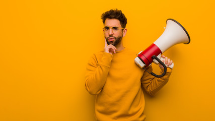 Young man holding a megaphone doubting and confused - obrazy, fototapety, plakaty
