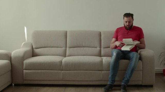 Caucasian beard man sitts on a sofa inside a room and reading a book. He doesnt know where to start the reading again and smilles