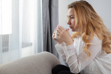 Caucasian blonde woman sitting on a sofa drinking her morning coffee in front of a window, casual and relaxed