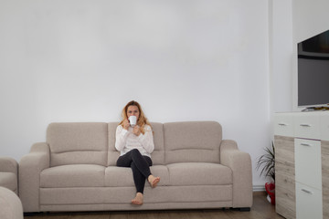 Obraz premium Caucasian blonde woman sitting on a sofa drinking her morning coffee, casual and relaxed