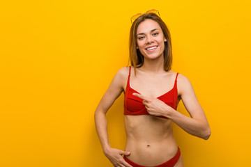 Young caucasian woman wearing bikini and sunglasses smiling and pointing aside, showing something at blank space.