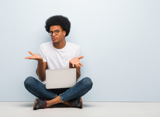 Young black man sitting on the floor with a laptop confused and doubtful - obrazy, fototapety, plakaty