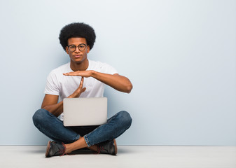 Young black man sitting on the floor with a laptop doing a timeout gesture