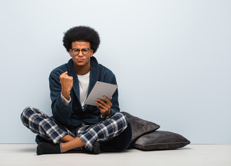 Young black man sitting on his house and holding his tablet showing fist to front, angry expression