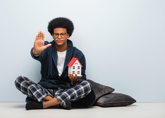 Young black man holding a house model sitting on the floor putting hand in front