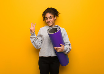 Young fitness black woman showing number three. Holding a mat.